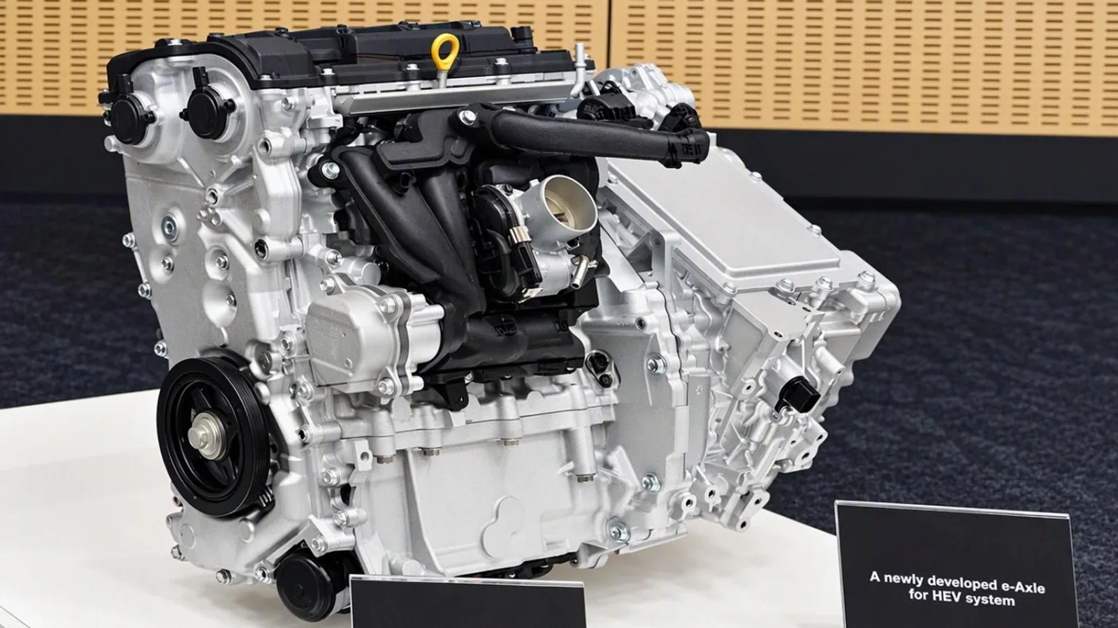 Motor Toyota hibrid de 1,5 litri și patru cilindri, expus pe un suport alb, fotografiat frontal într-un spațiu cu perete bej în spate. Motorul este de culoare argintie cu componente negre, iar în față se vede o placă informativă neagră.