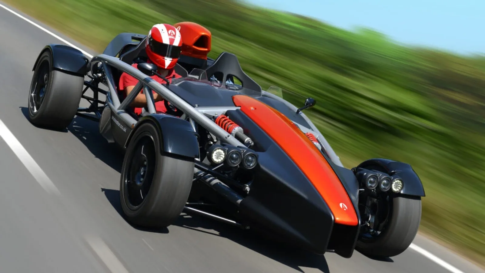 Mașină sport Ariel Atom 4 neagră cu dungi portocalii, văzută din față într-o curba pe drum asfaltat, fundal defocalizat verde cu copaci, accent pe structura tubulară și designul aerodynamic deschis.