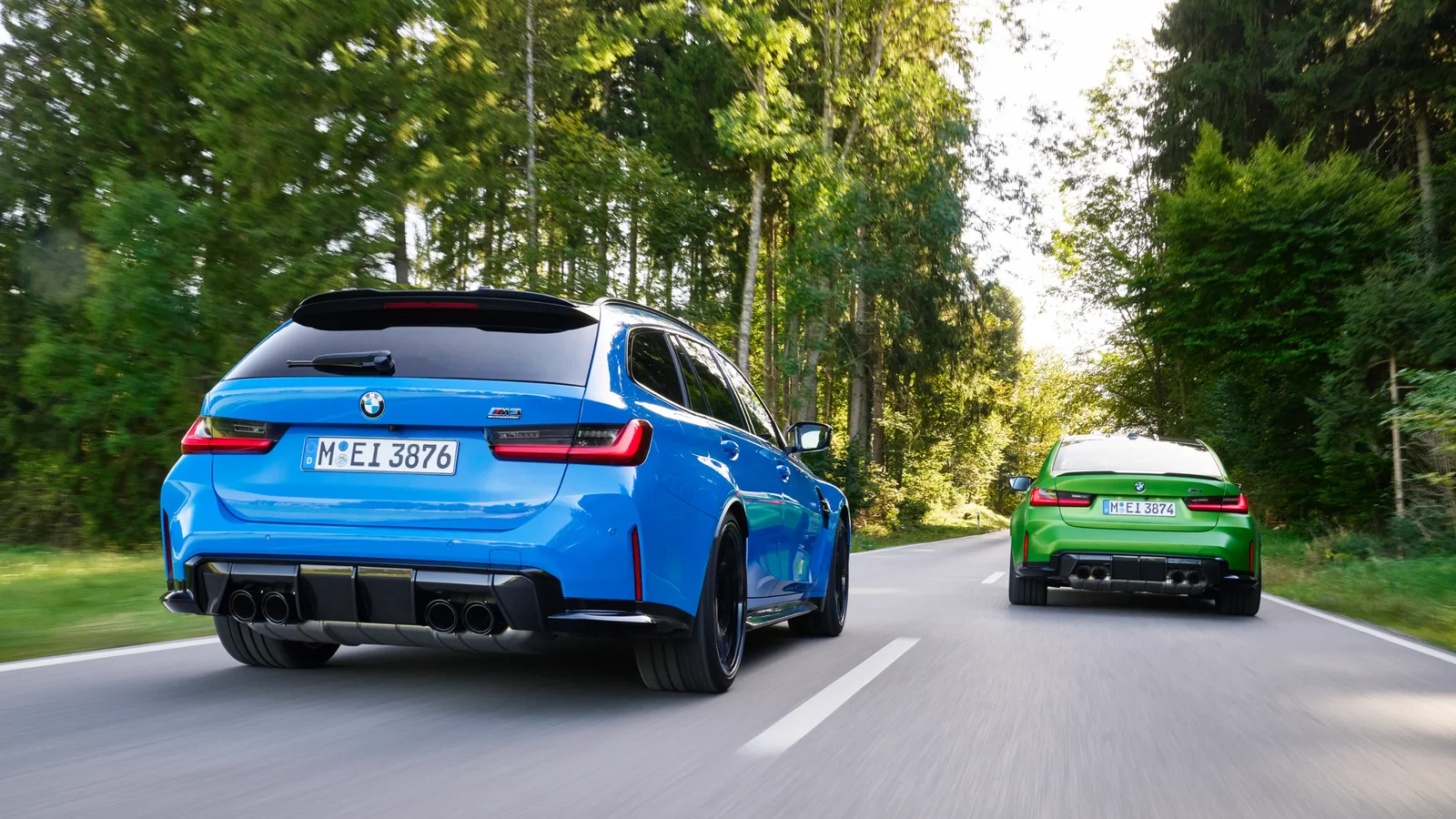BMW M3 Touring albastru și BMW M3 Touring verde în mers pe un drum asfaltat în pădure, văzute din spate la un unghi jos, în condiții de lumină naturală.