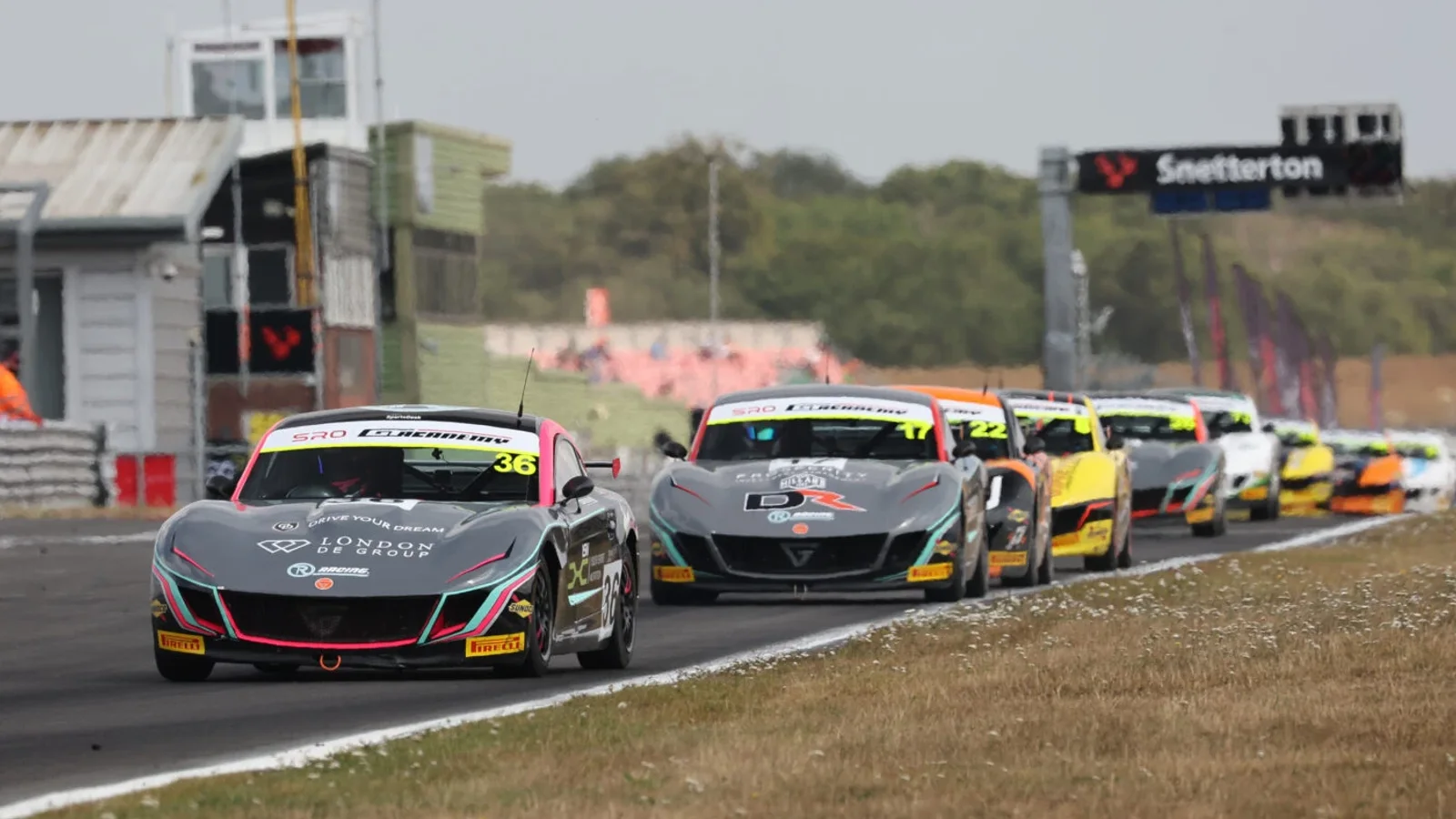 Mai multe mașini de curse Ginetta Junior, aliniate pe circuitul Snetterton, parcurg linia dreaptă cu vegetație și clădiri în fundal, fotografiate dintr-un unghi lateral, jos, sub cer parțial noros.