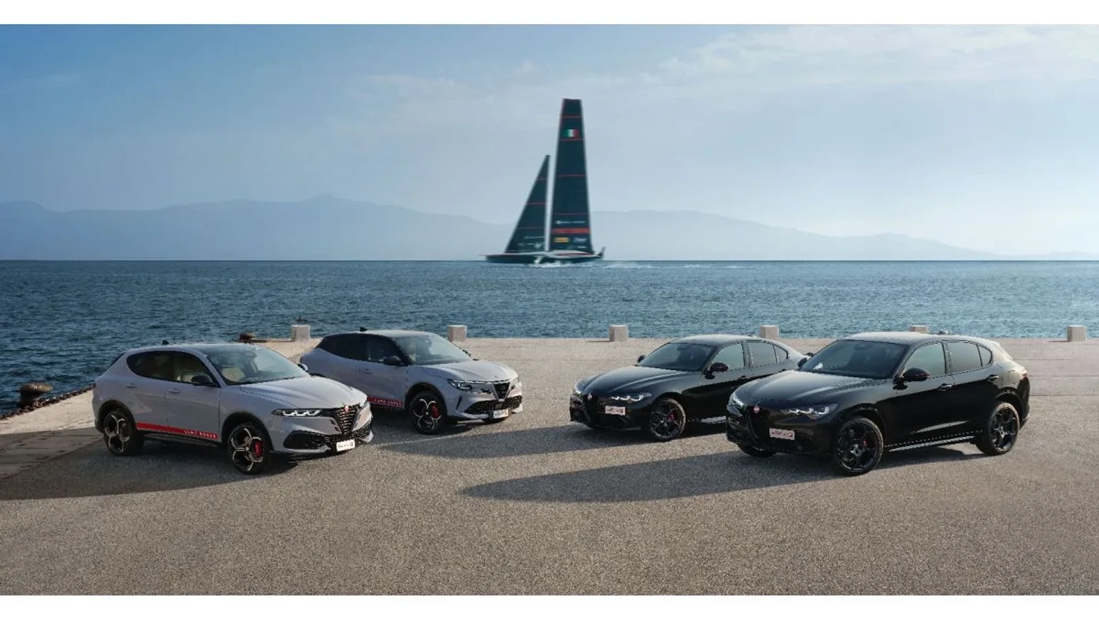 Modele Alfa Romeo Edizione Luna Rossa aliniate pe malul mării, cu velierul Luna Rossa în fundal.