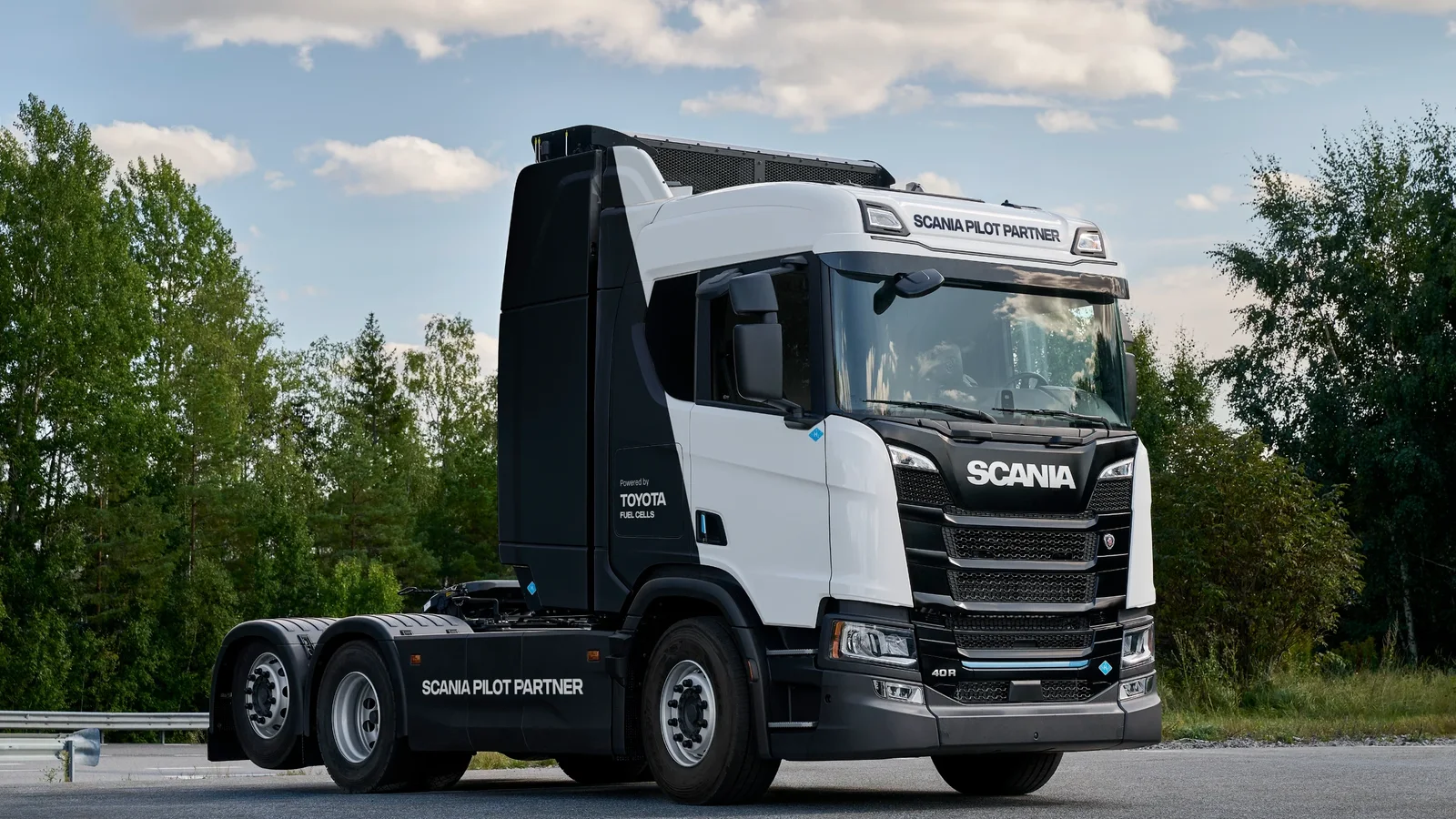 Camion Scania alb pe șosea, văzut din față și lateral, cu copaci verzi și cer parțial înnorat în fundal.