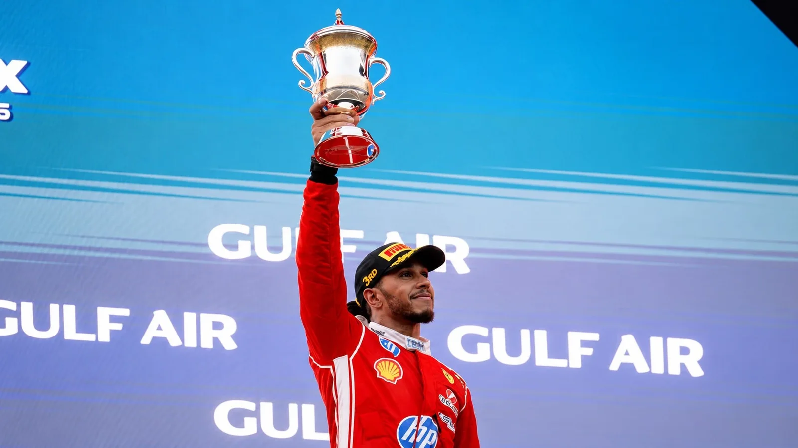 Lewis Hamilton în echipament Ferrari roșu ridică trofeul pentru locul al treilea pe podiumul Marelui Premiu al Bahrainului.