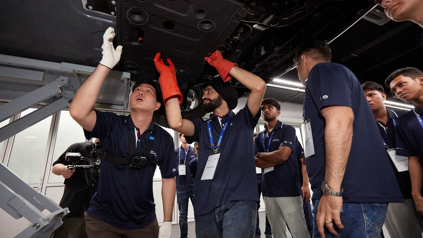 Cursanți Hyundai inspectează componentele de sub un automobil ridicat pe elevator, în timpul unui program de pregătire tehnică.