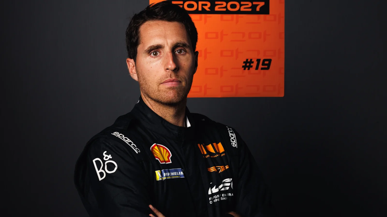 Portret cu Dani Juncadella în combinezon negru de curse cu sigle colorate, pe fundal întunecat cu un panou portocaliu pe perete pe care scrie „SIGNED FOR 2027” și numărul 19, privire serioasă, brațele încrucișate, lumină din față.