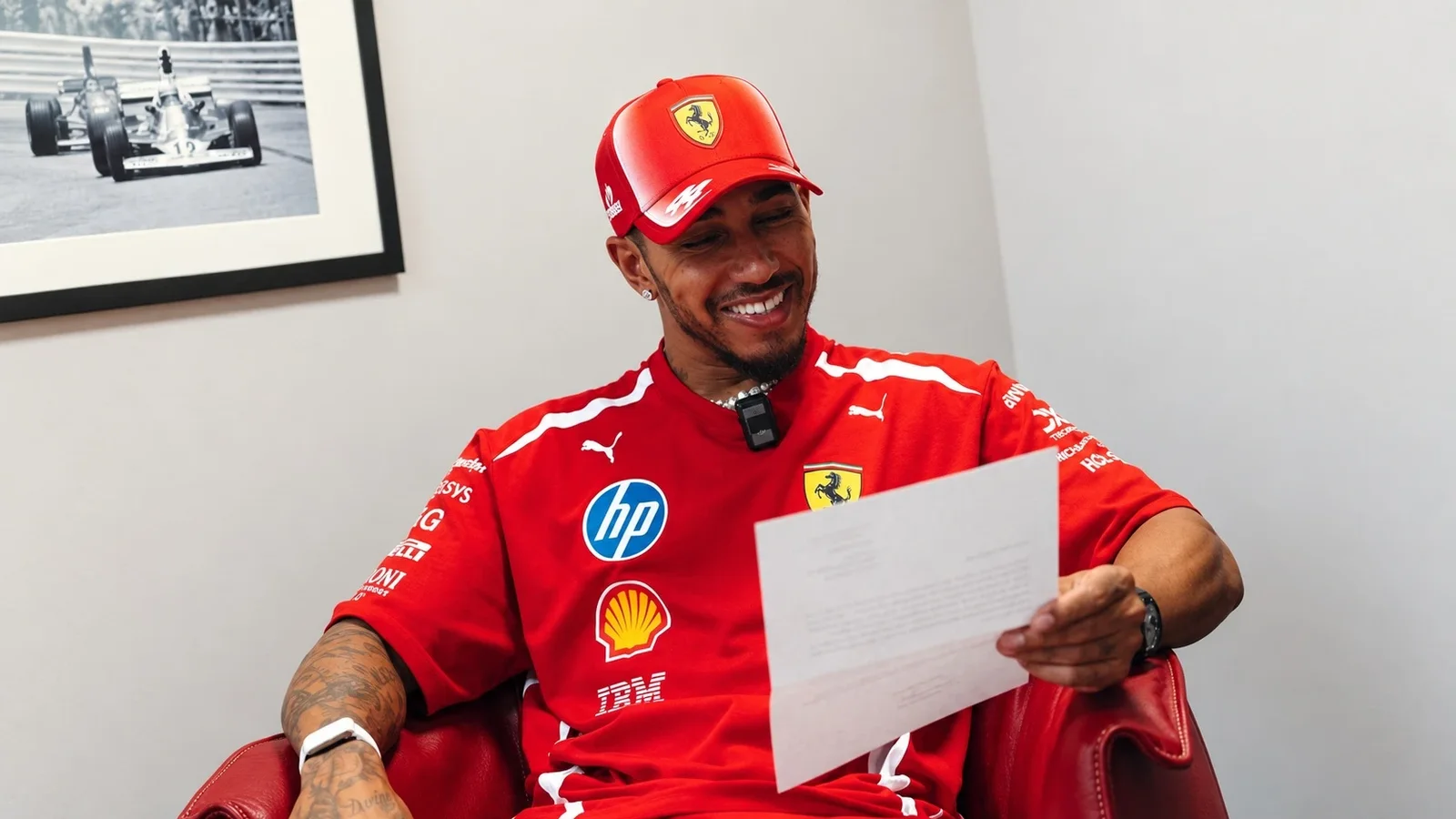 Lewis Hamilton, îmbrăcat în echipamentul Ferrari, citește un document într-un fotoliu roșu.
