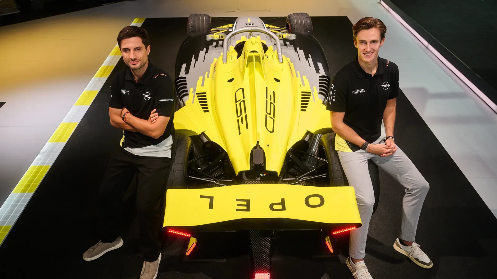 Monopostul electric Opel GSE 27FE galben cu negru și argintiu, văzut de sus din față, expus pe podium interior. În stânga și dreapta sunt doi tineri îmbrăcați în tricouri negre și pantaloni deschiși, stând pe genunchi, zâmbind lângă mașină. Fundal întunecat cu iluminare direcționată.