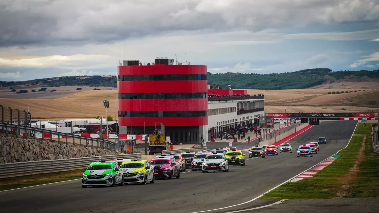Mai multe mașini de curse Renault Clio Cup în culori verde, alb și roz, în accelerare pe pistă la o curbă, cu un turn roșu și negru și peisaj rural în fundal sub un cer noros.