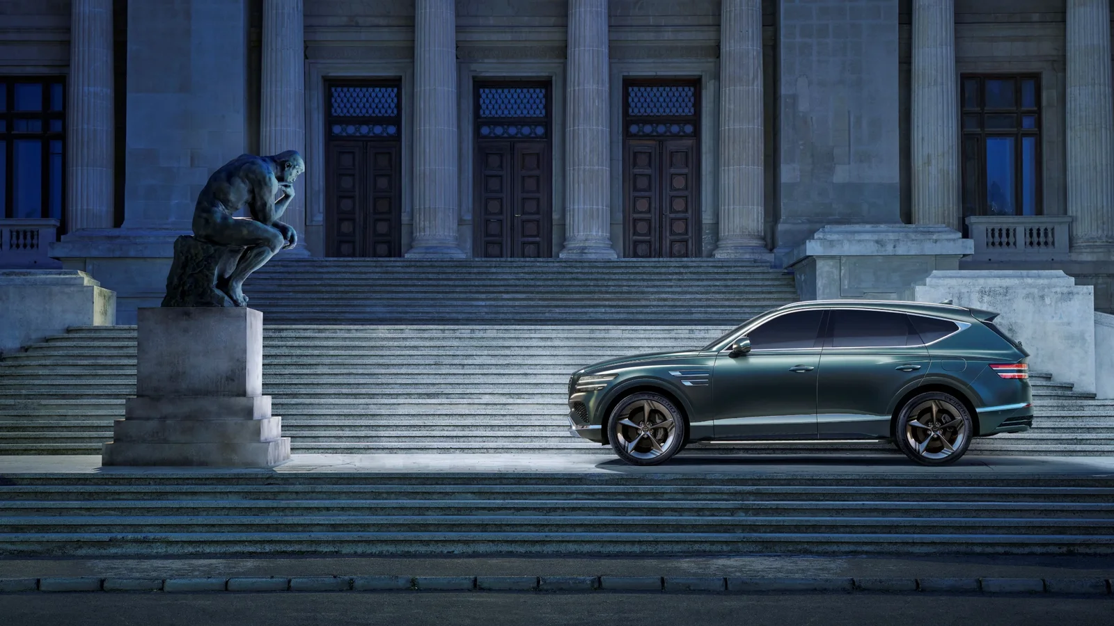 SUV Genesis GV80 Hybrid gri închis parcat pe trepte largi, cu clădire monumentală cu coloane în fundal, fotografie realizată din lateral, în lumină albastră ambientală de seară.