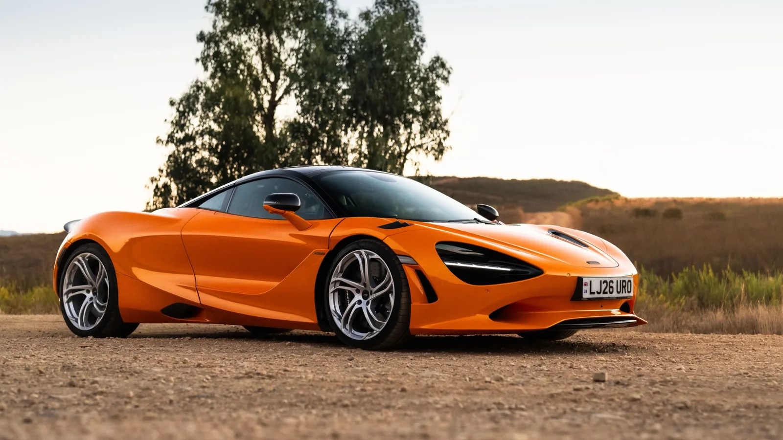 McLaren 750S portocaliu, fotografiat din față și lateral pe un drum de țară, la apus