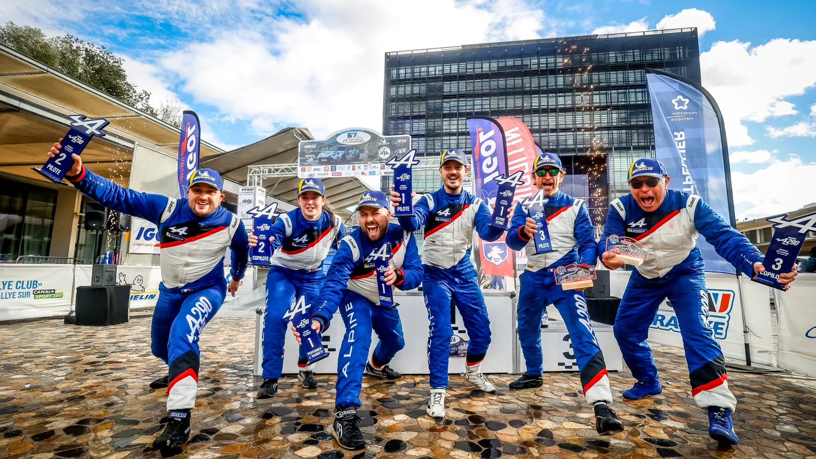 Echipajul Fun Meca Sport, format din cinci bărbați în combinezoane albastre și alb-roșii, sărbătorește victorie în fața clădirii alpine, pe o piață cu pavele umede, sub cer parțial înnorat. Drapele și bannere Alpine sunt vizibile în fundal.