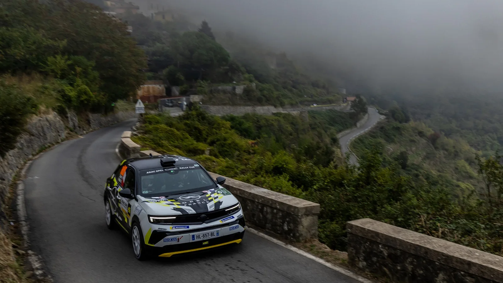 Opel Mokka GSE Rally în mers pe un drum montan îngust, cu parapet de piatră, vizibil parțial sub ceață. Mașina are un decor grafic alb, negru și verde și este fotografiată din față, ușor lateral, pe un timp înnorat.