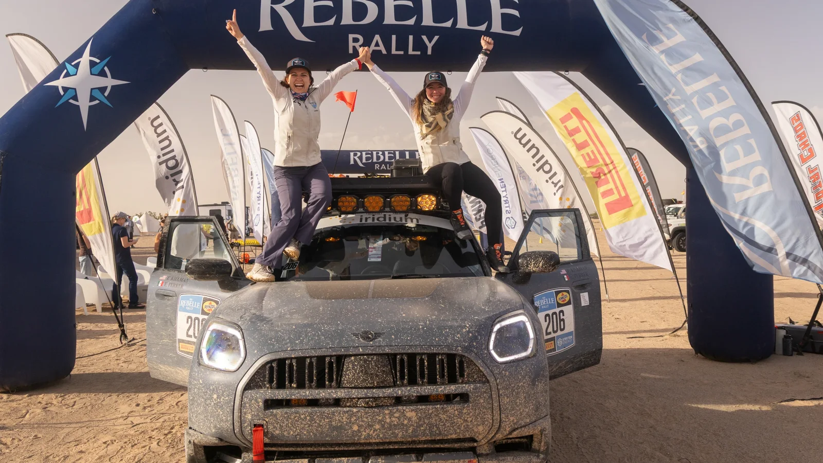 MINI Countryman Untamed Edition acoperit cu noroi, oprit sub un arc albastru de start/sosire La Rebelle Rally, cu două femei în picioare pe plafon, ridicând brațele în semn de victorie, zgomot de fundal deșertic și drapeluri laterale.