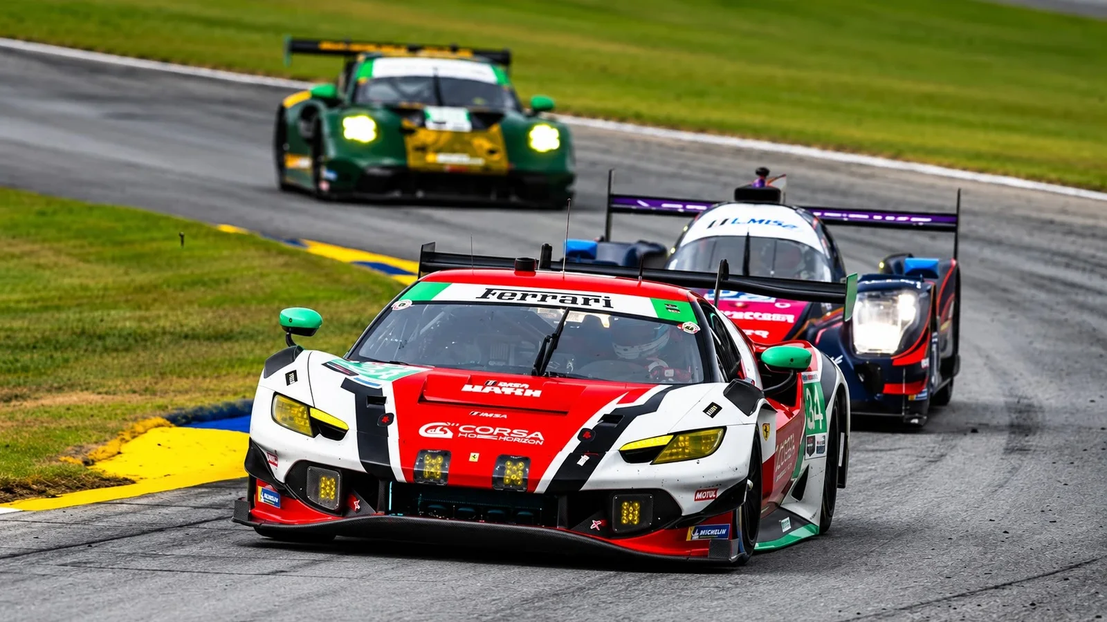 Ferrari 296 GT3 Evo în culorile alb, roșu și verde, rulând pe pistă udă la Petit Le Mans, urmat de alte mașini de curse.