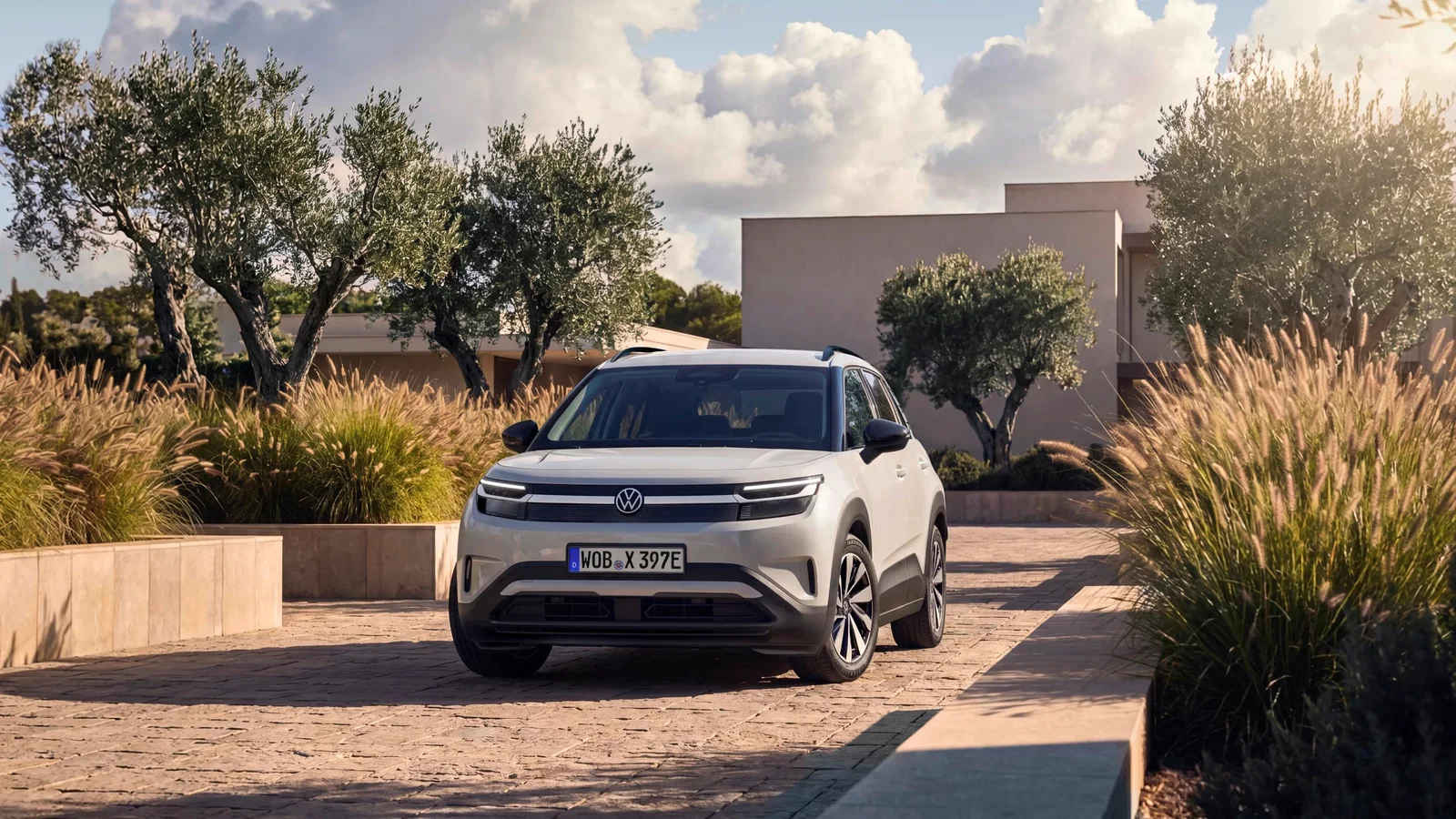 Volkswagen ID. Cross alb, văzut din față pe o alee pietruită, înconjurat de vegetație joasă și copaci, sub un cer parțial noros, lumină caldă de apus, într-un cadru urban modern.