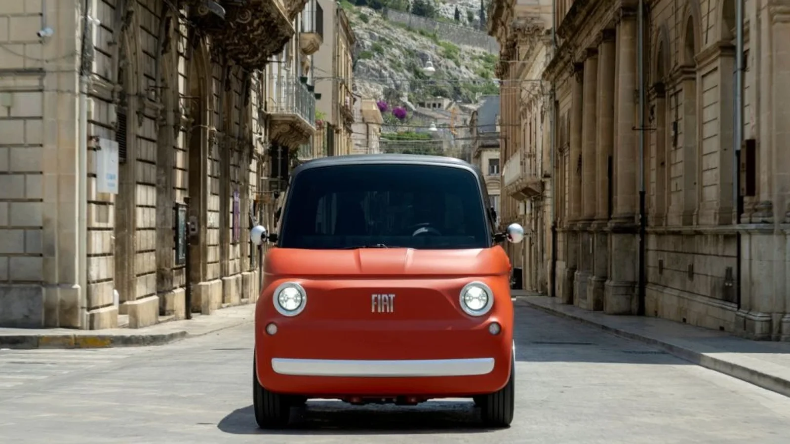 Fiat electric portocaliu, văzut din față pe o stradă îngustă dintr-un oraș istoric.