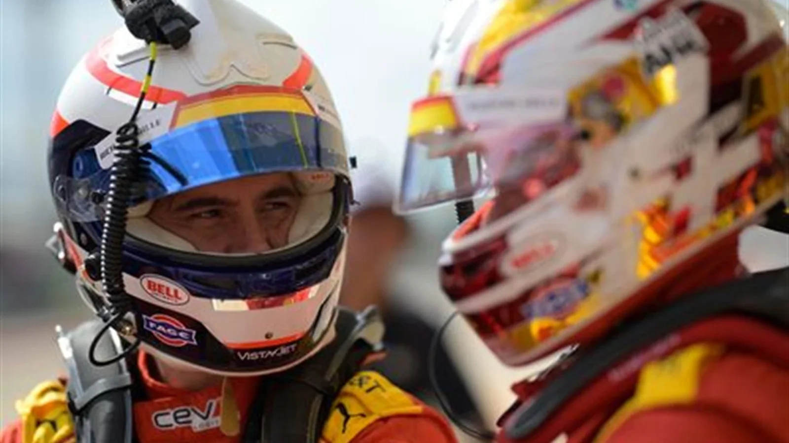 Doi piloți de curse în combinezoane roșii și căști discută față în față înaintea unei sesiuni pe circuit.