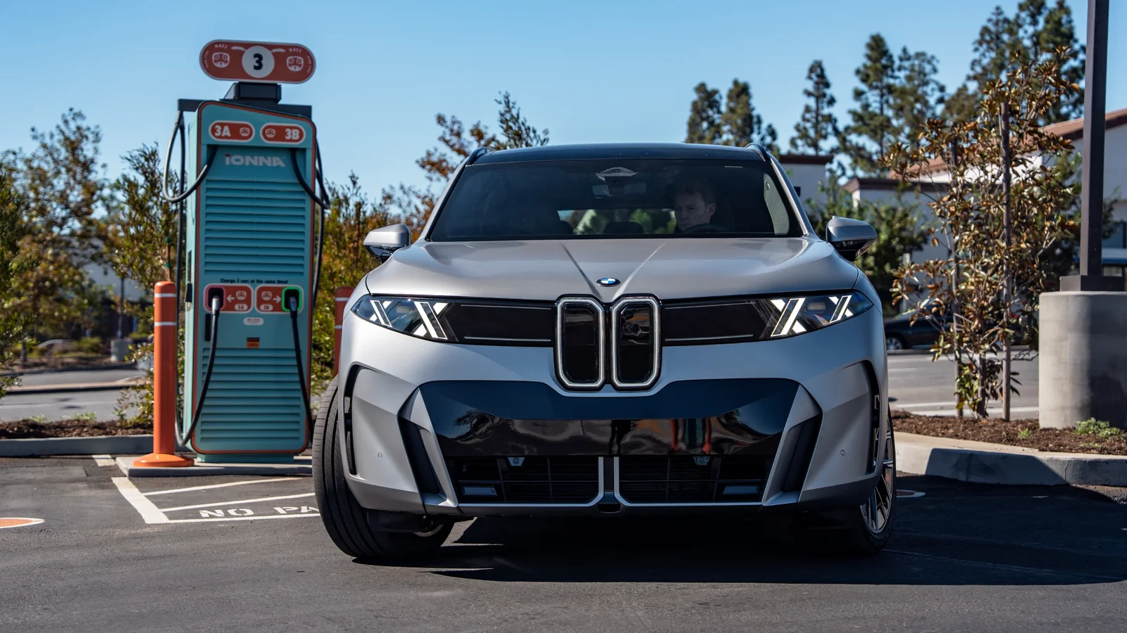 BMW iX5 60 xDrive, față, culoare argintie, fotografiat frontal în parcare lângă stație de încărcare electrică vintage, cer senin, lumină naturală, în sudul Californiei.