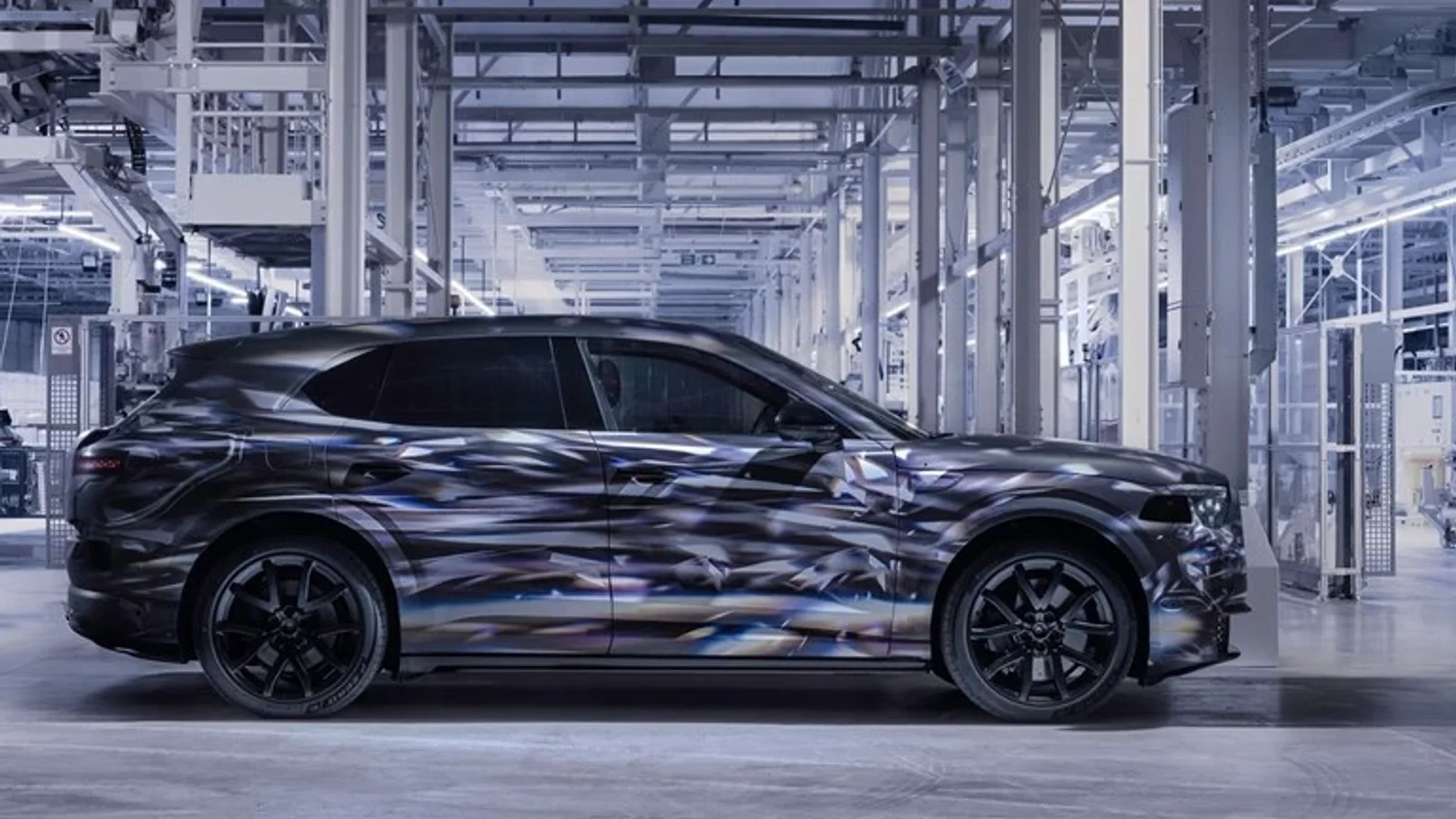 Bentley Torcal prototip electric cu camuflaj gri-negru, văzut din profil într-un spațiu industrial luminos, cu echipamente și structuri metalice în fundal, interiorul halei având o cromatică preponderent argintie și albă.