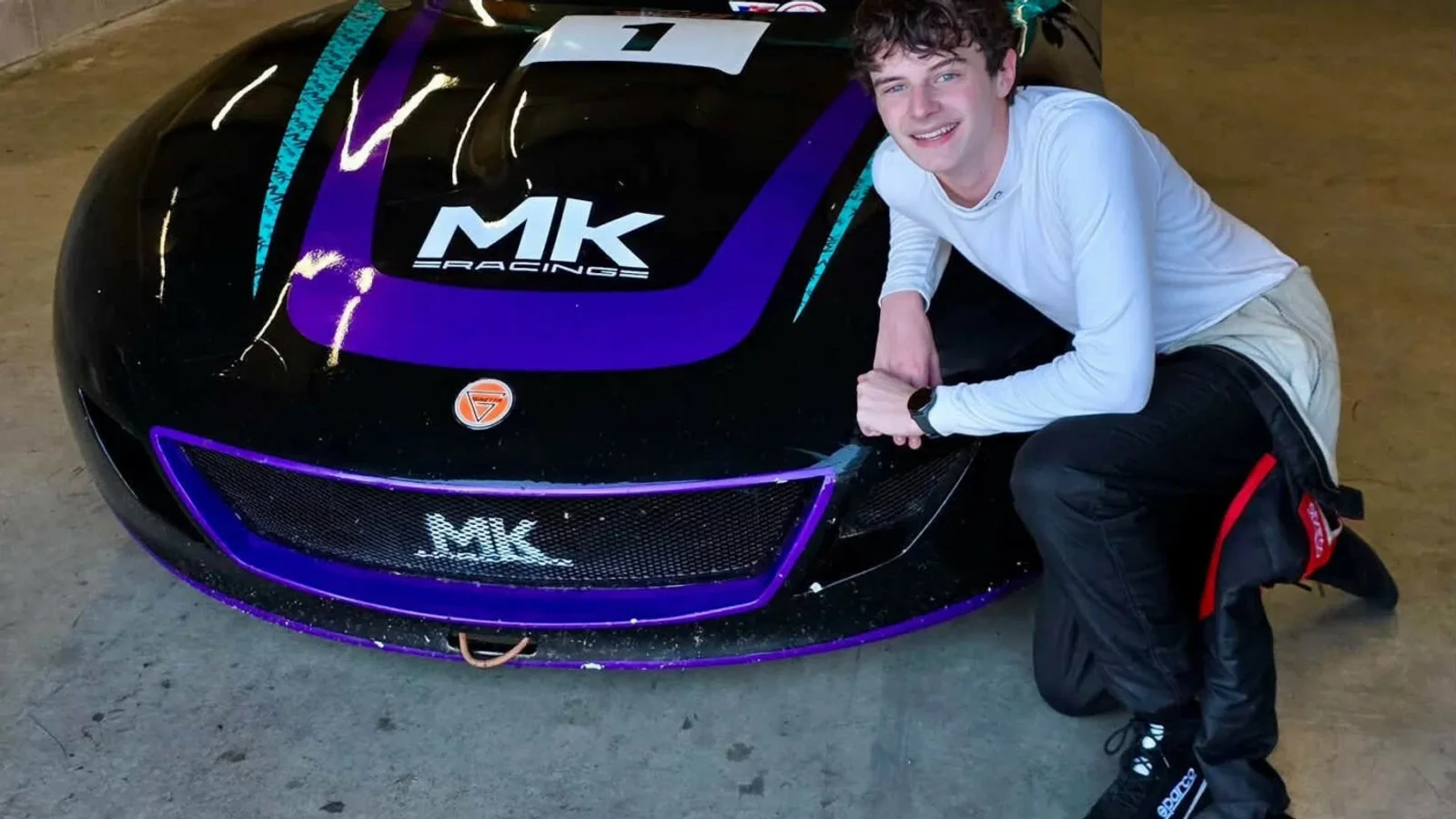 Tânăr pilot îmbrăcat în combinezon de curse alb și negru, așezat pe genunchi lângă un automobil de curse Ginetta, culoare neagră cu elemente mov și logo MK Racing pe capotă, fotografie realizată într-un garaj.