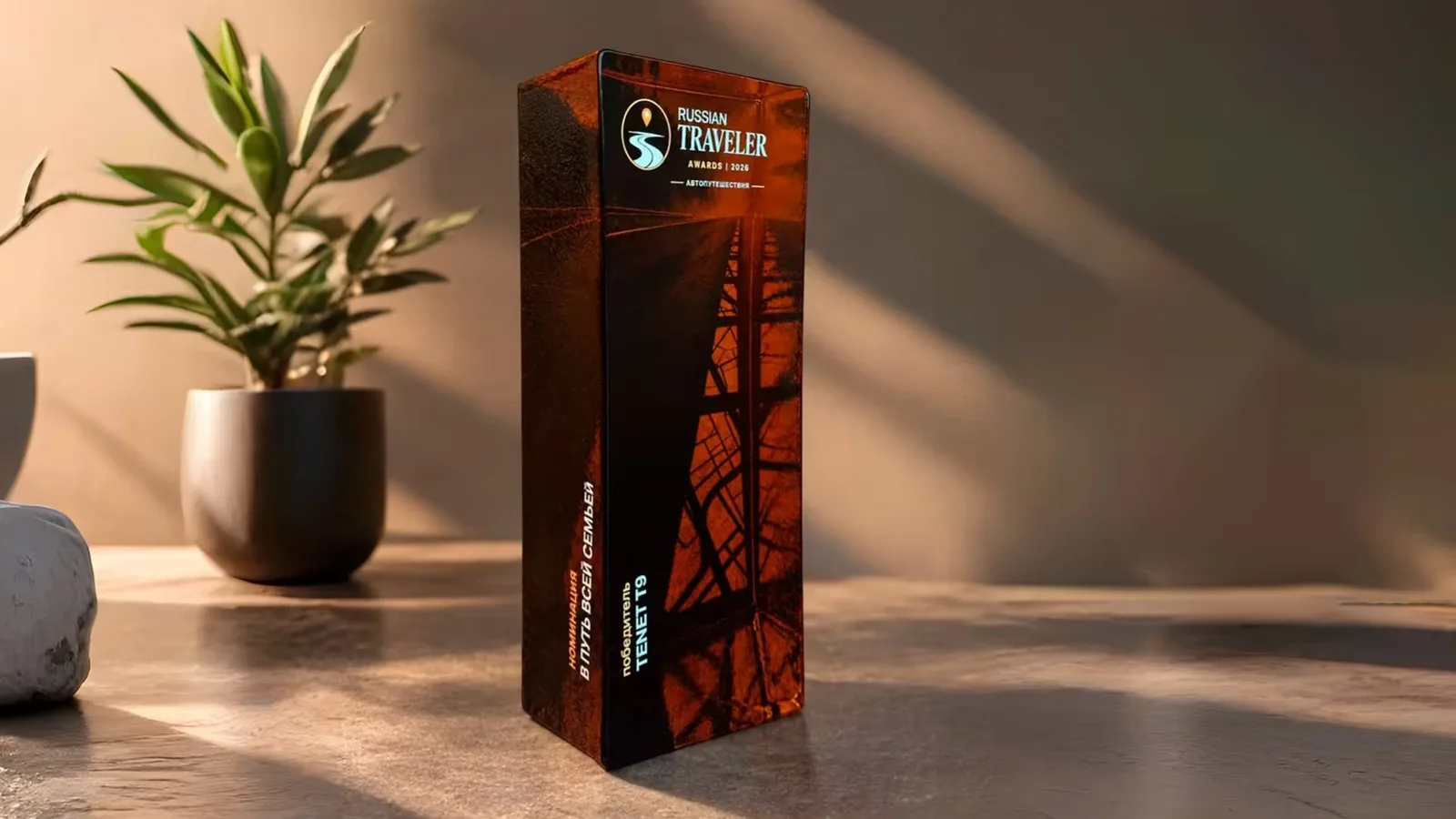 Trofeu premiu Russian Traveler Awards expus pe o masă, într-o cameră luminată natural. Trofeul are un design vertical dreptunghiular, cu detalii în nuanțe de maro și negru. În fundal, o plantă verde în ghiveci și un perete bej.