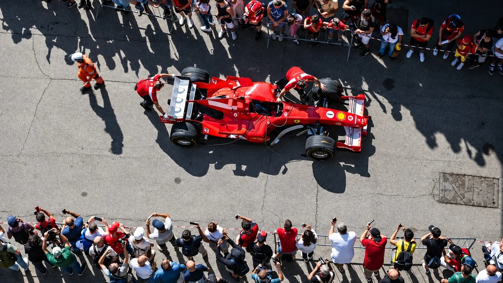 Monopost Ferrari roșu de Formula 1, văzut de sus în paddockul Mugello, înconjurat de membri ai echipei și spectatori.