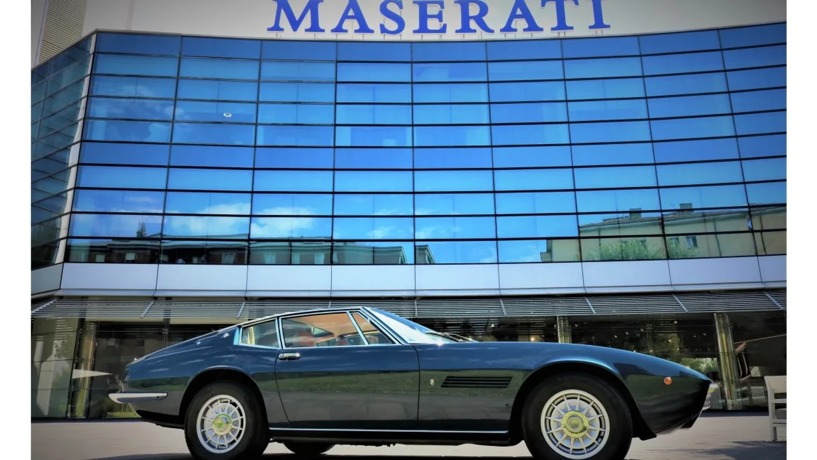 Vehicul Maserati Ghibli SS Coupé din anii ’70, vopsit în albastru închis, fotografiat lateral în fața unei clădiri moderne cu fațadă din sticlă, cu logo-ul Maserati vizibil deasupra intrării, sub o lumină naturală, înplan deschis.