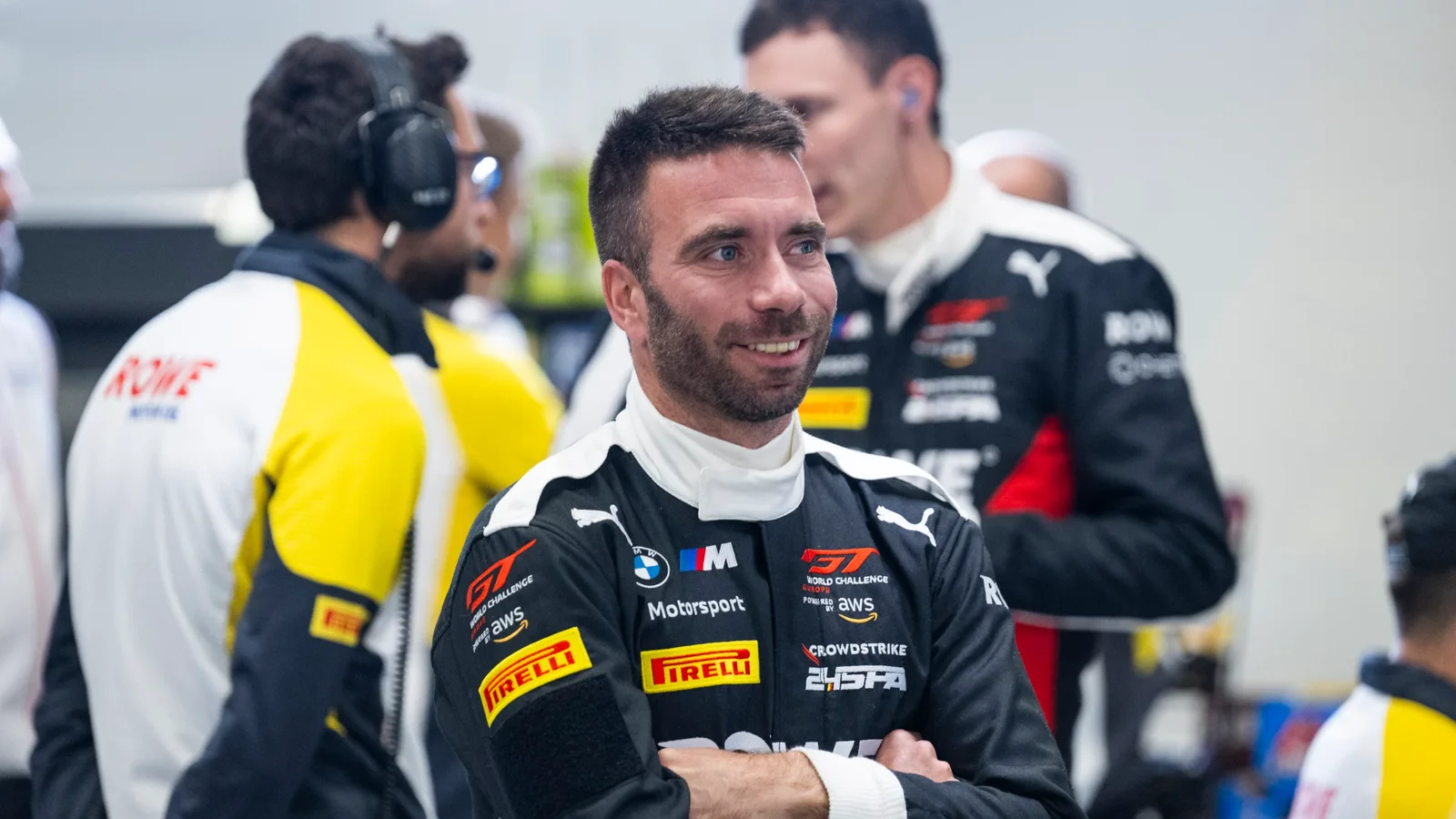Philipp Eng în combinezon negru cu logo-uri BMW M Motorsport și Pirelli, zâmbind cu brațele încrucișate într-un garaj de curse, oameni în fundal cu echipament de echipă. Imagine din cadrul unui eveniment motorsport.