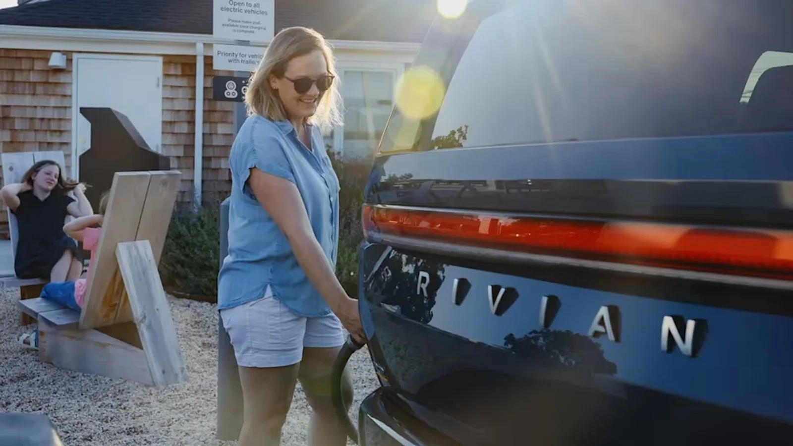Femeie încărcând un SUV Rivian la o stație publică de încărcare electrică, în lumina apusului.