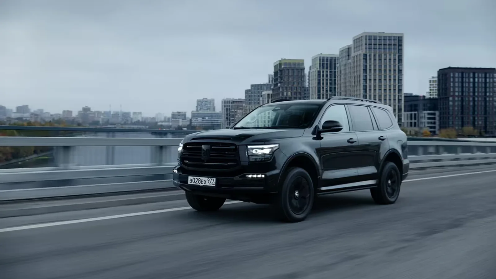 SUV negru TANK 500 în mișcare pe un pod urban, cu clădiri înalte și cer noros pe fundal, vedere laterală dreaptă la nivel mediu.