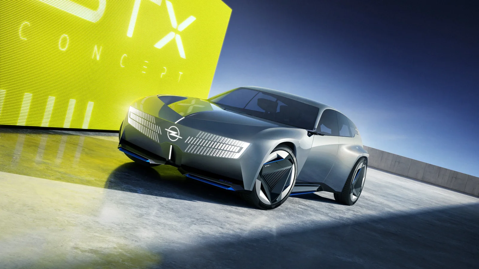 SUV concept Opel STX gri metalizat, fotografiat din unghi frontal stânga, cu faruri LED și jante mari, pe fundal albastru și galben cu text STX Concept vizibil parțial.