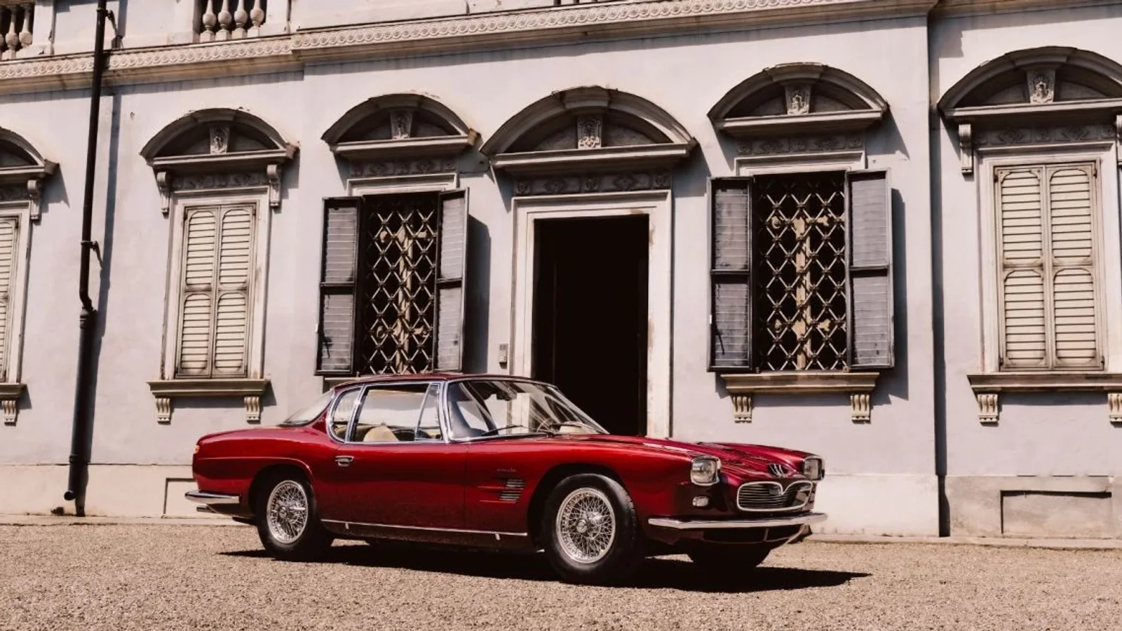 Maserati 5000 GT Frua roșu, parcat în fața unei clădiri istorice cu ferestre înalte.