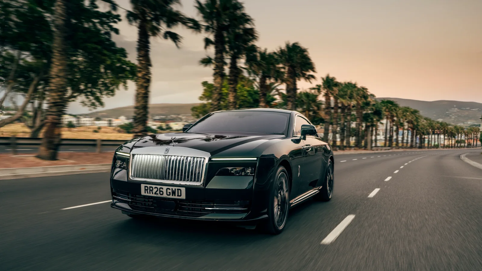 Rolls-Royce Spectre Series II negru în mișcare pe un drum asfaltat flancat de palmieri, fotografiat din față și unghi jos, pe timp de zi, cu cer senin și munți în fundal.