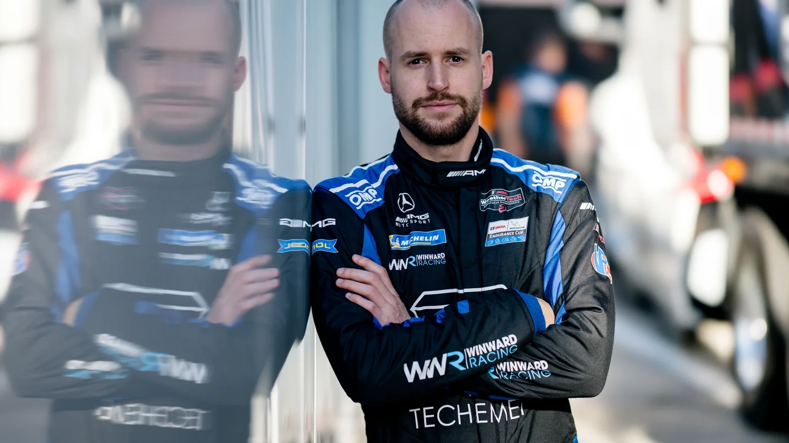 Portret al pilotului Kimi Antonelli în combinezon albastru închis cu brațele încrucișate, reflectat pe o suprafață lucioasă, fundal neclar cu elemente urbane.