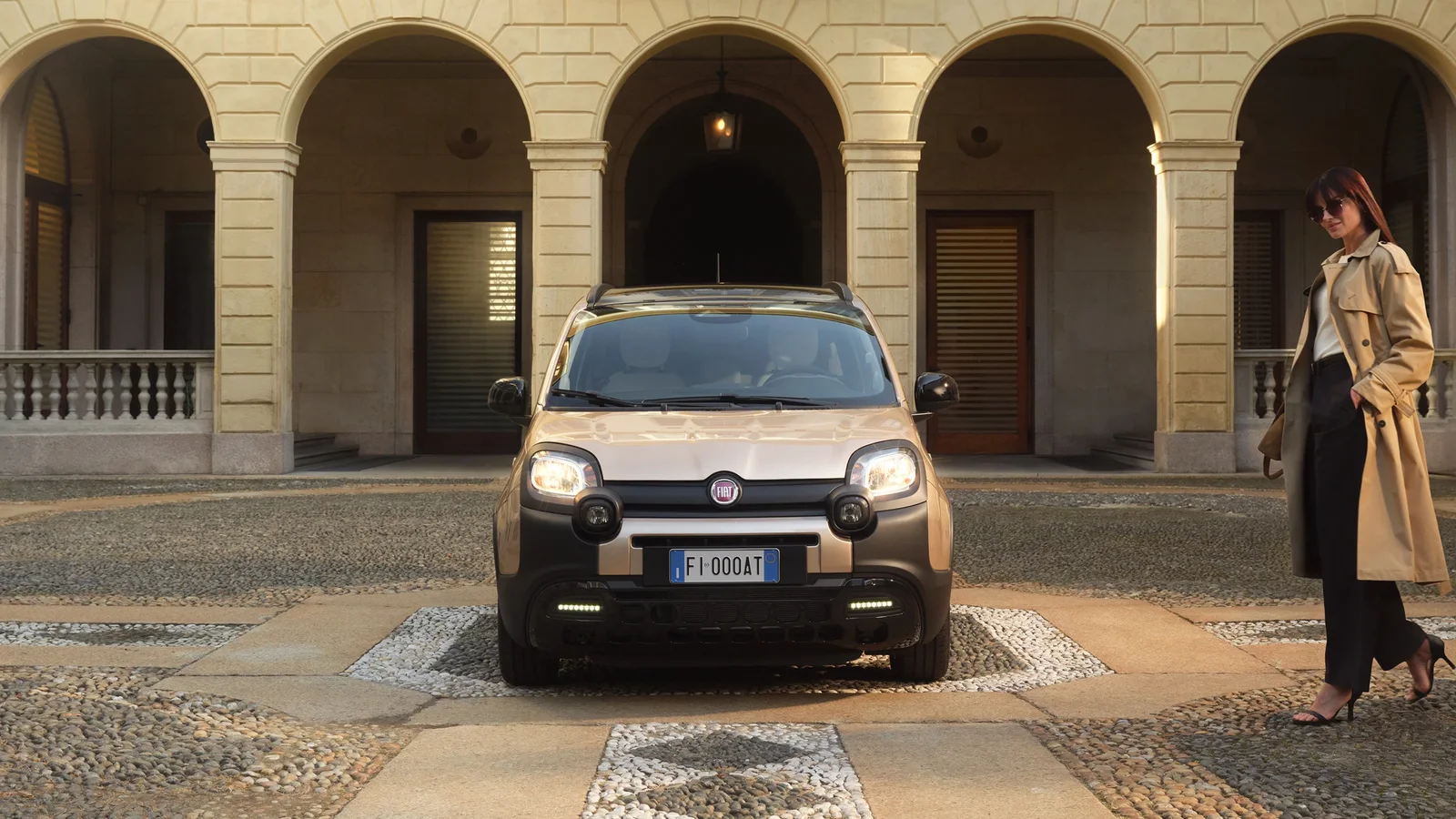 Fiat Pandina Tributo Autobianchi parcat frontal într-un spațiu urban cu pavaj, clădire istorică cu arcade în fundal. Mașina are vopsea Platino deschisă și elemente negre vizibile, iar o femeie poartă o haină bej în dreapta imaginii.