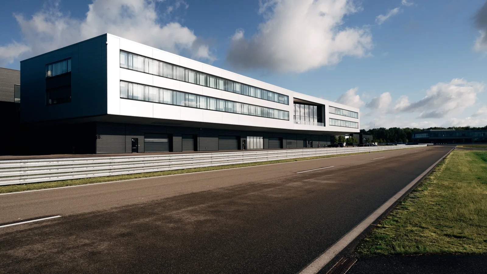 Clădire modernă cu fațadă albă și ferestre largi, situată lângă un circuit de curse asfaltat, pe fundal cer albastru cu nori albi, la lumina zilei; sediul Audi Formula Racing din Neuburg an der Donau.