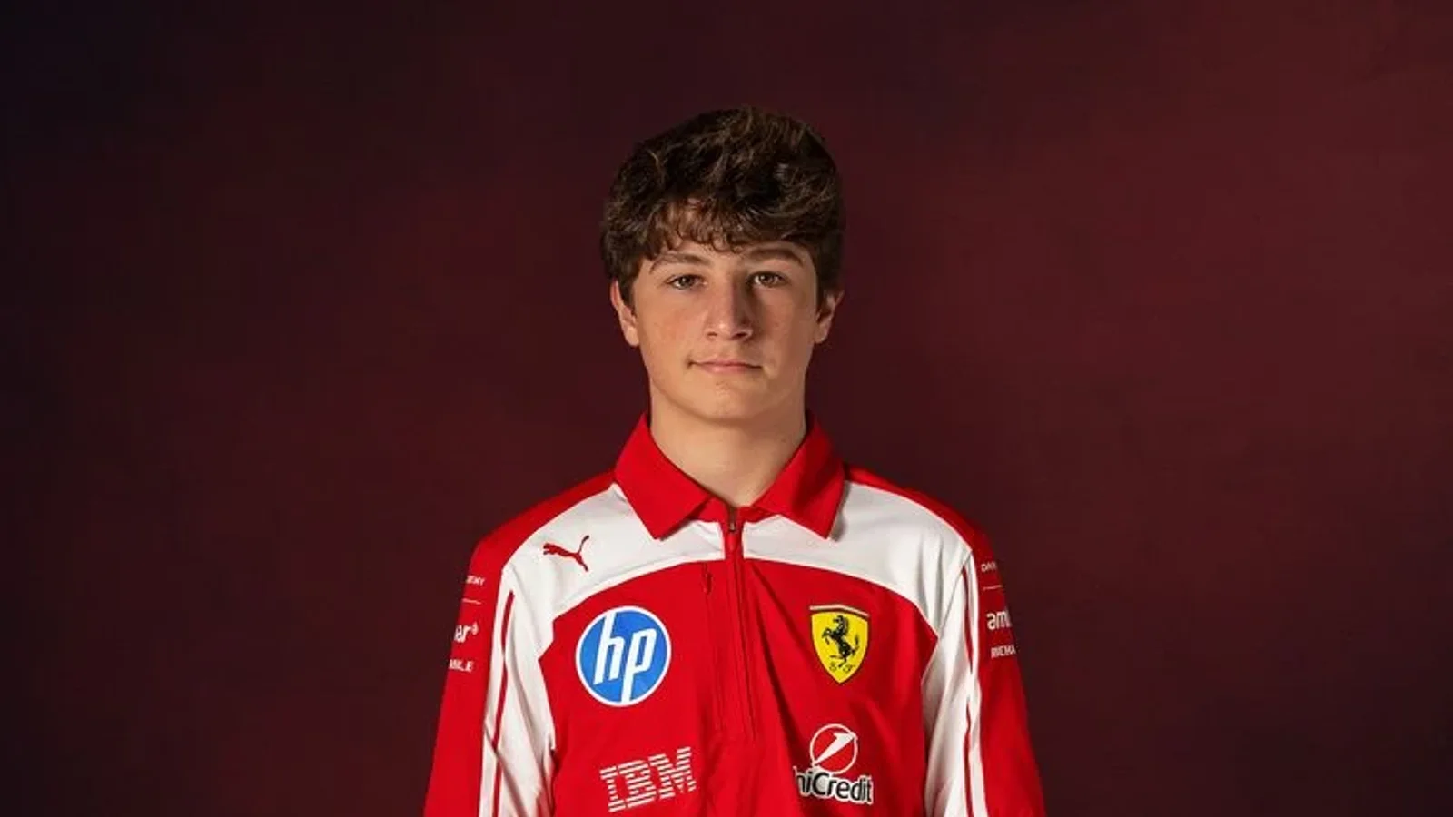 Portret frontal al lui Niccolò Maccagnani, pilot Ferrari, purtând echipament roșu cu logo-uri Ferrari și sponsori, pe fundal de culoare bordo.