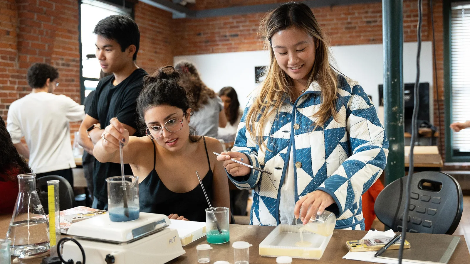 Tineri cercetători și studenți într-un laborator de design industrial, lucrând la prototipuri și experimente cu echipamente de laborator pe o masă. În fundal, ferestre mari și pereți din cărămidă roșie, iluminare naturală și artificială.