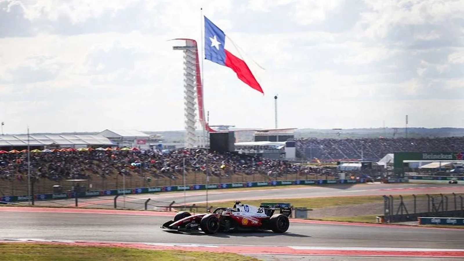 Monopost Ferrari în viteză pe circuitul Circuit of the Americas, Austin, pe un viraj cu bordură roșie, sub un cer parțial senin; în fundal, tribune pline și drapelul statului Texas fluturând pe un catarg înalt.