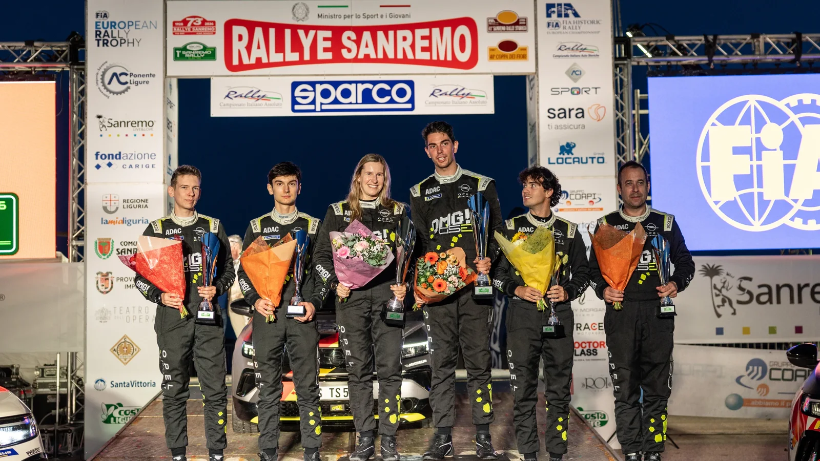 Șase piloți de raliu, îmbrăcați în costume de curse negre, stau pe podiumul Raliului Sanremo, ținând trofee și flori. În spatele lor sunt bannere cu logo-ul „Rallye Sanremo” și simboluri FIA. Fotografie realizată frontal, pe timp de zi, la un eveniment oficial.