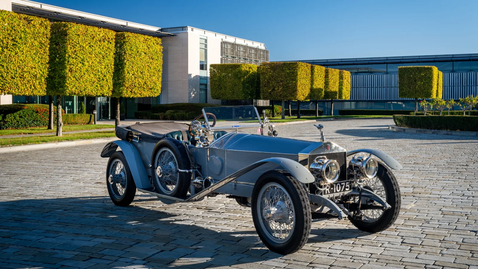 Rolls-Royce Silver Ghost din 1911, șasiul 1701, mașină clasică decapotabilă argintie parcată pe o piațetă cu pavaj, în fața unor clădiri moderne cu fațade acoperite parțial de vegetație, fotografiată din unghi lateral-jos, prim-plan spre partea frontală.