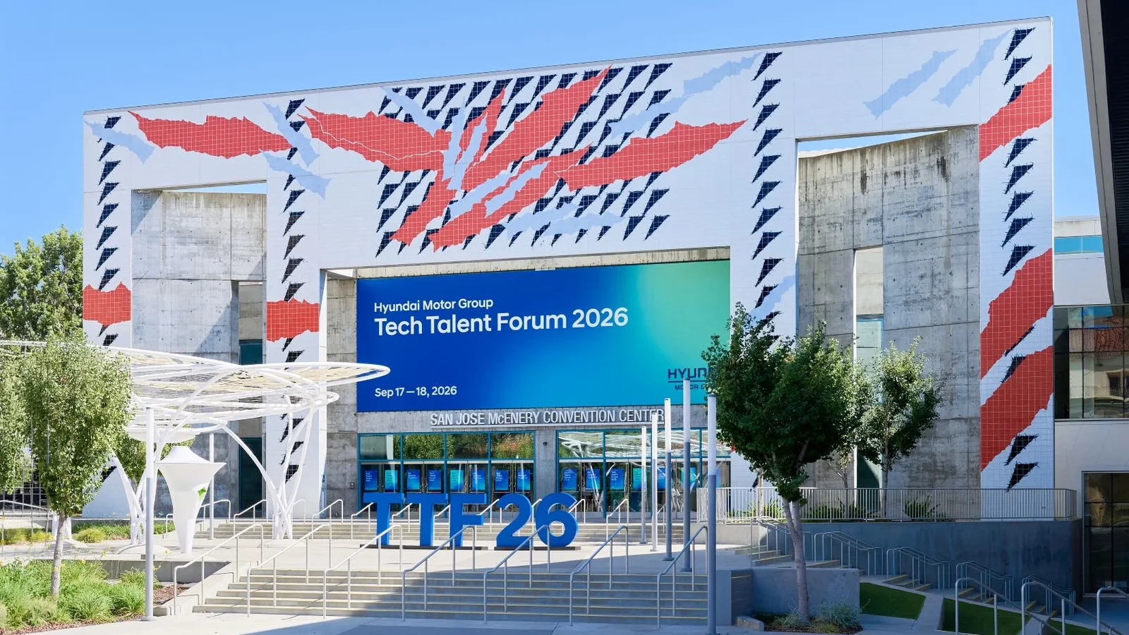 Fațadă modernă a centrului de convenții McEnery din San Jose decorată cu un design geometric roșu și albastru, în față afiș digital cu text despre Tech Talent Forum 2026, decor ambiental luminos, vedere din față pe timp de zi cu cer senin.