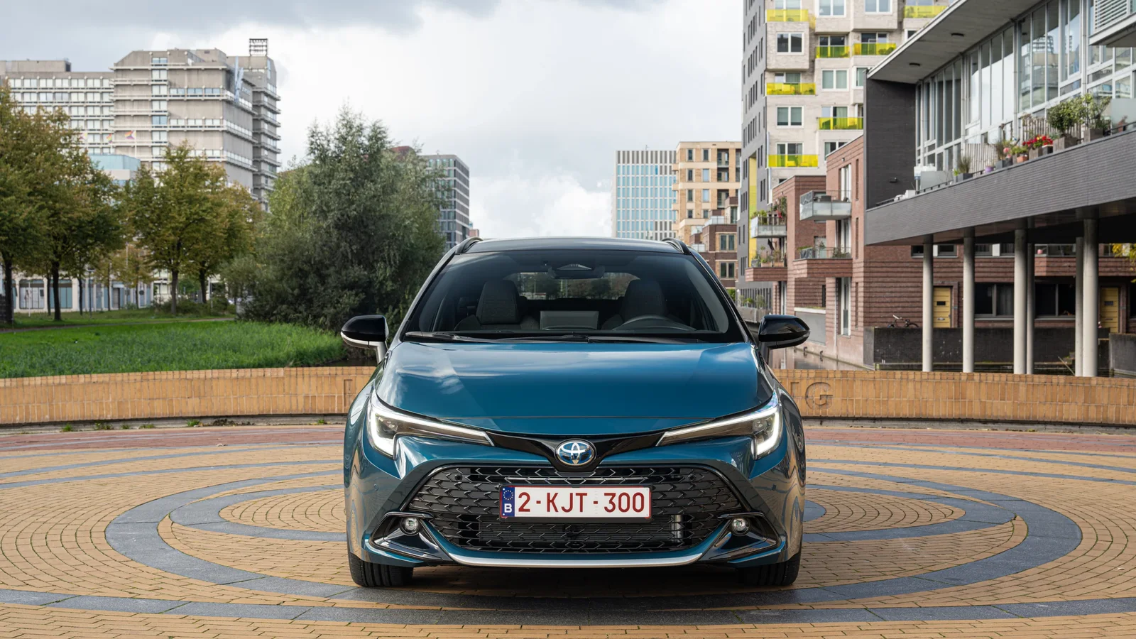 Toyota Corolla Hatchback albastru New Boreal Blue, văzută frontal pe o piațetă urbană cu clădiri și copaci în fundal, iluminare naturală, accent pe grila proeminentă și faruri LED subțiri.