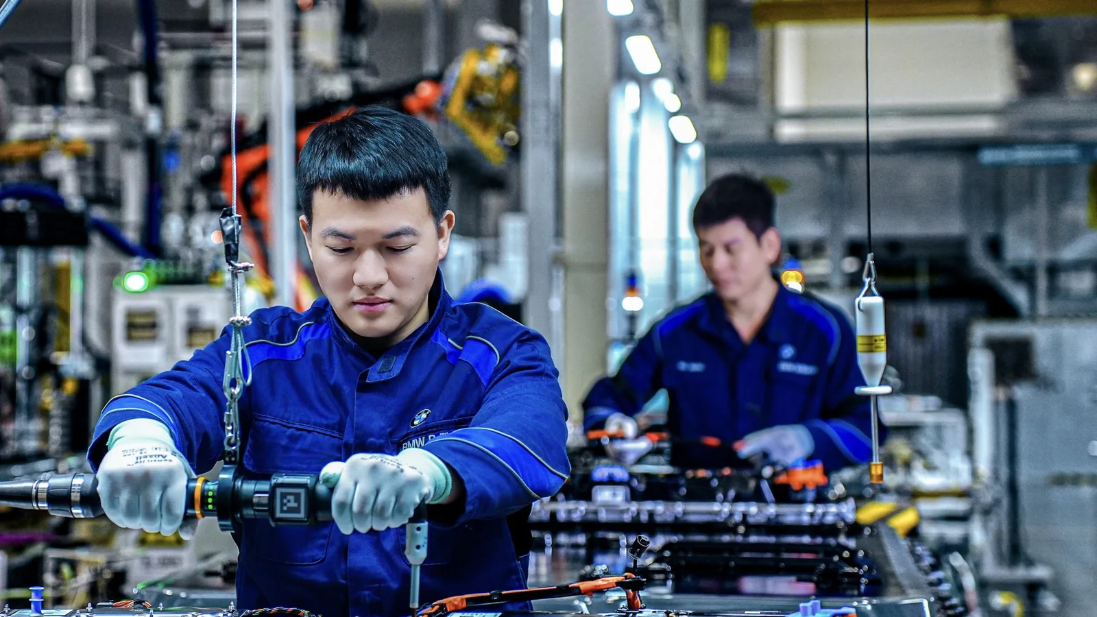 Doi muncitori asiatici în salopete albastre asamblează componente electrice pe o linie de producție în fabrica BMW din Shenyang, China. Foto surprinde în plan apropiat accesorizarea cu echipamente de lucru, cu hale mari și instalații industriale vizibile în fundal.