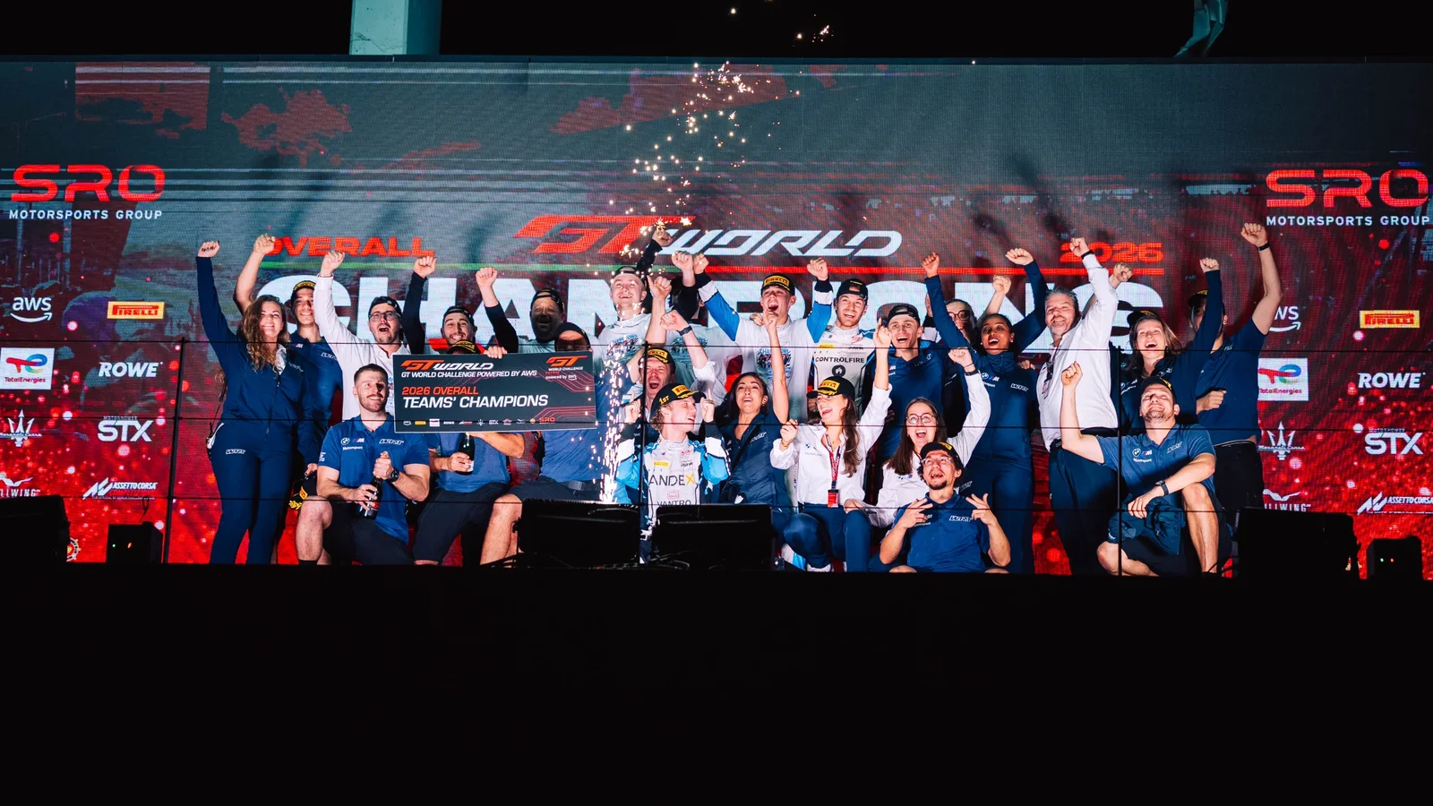 Echipajul Team WRT sărbătorește pe podium victoria în GT World Challenge, în fața unui fundal roșu cu logo-uri, în noapte, cu artificii și confetti. Toți membrii sunt în echipament sportiv, unii ridicând trofee și brațele în aer.