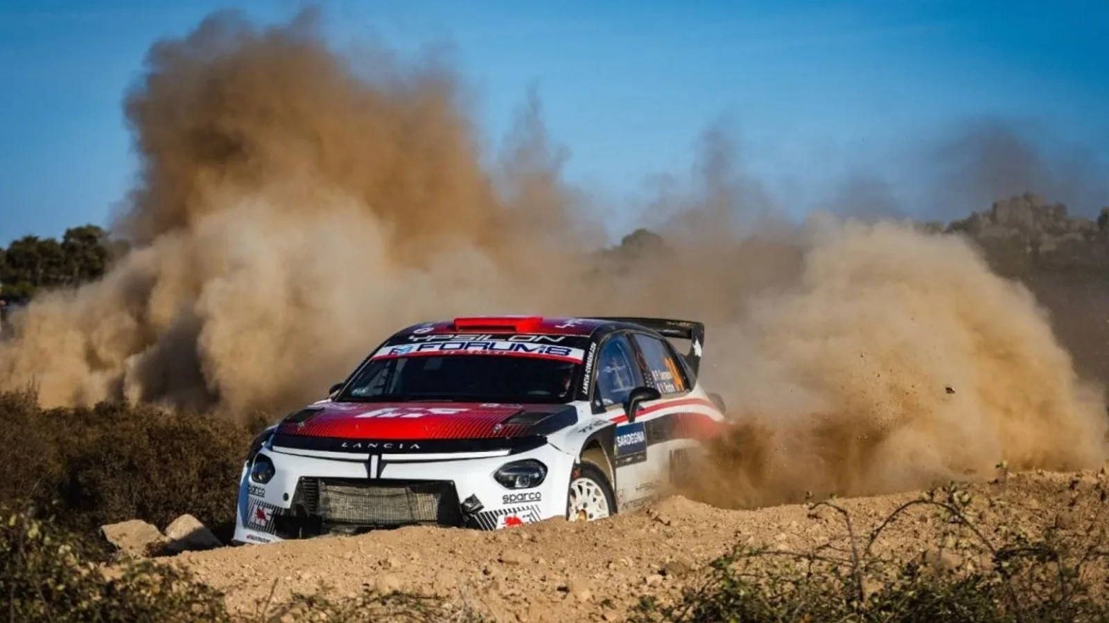 Lancia Ypsilon Rally2 HF Integrale în culorile alb, roșu și negru, rulând pe un traseu de macadam din Sardinia și ridicând un nor dens de praf.