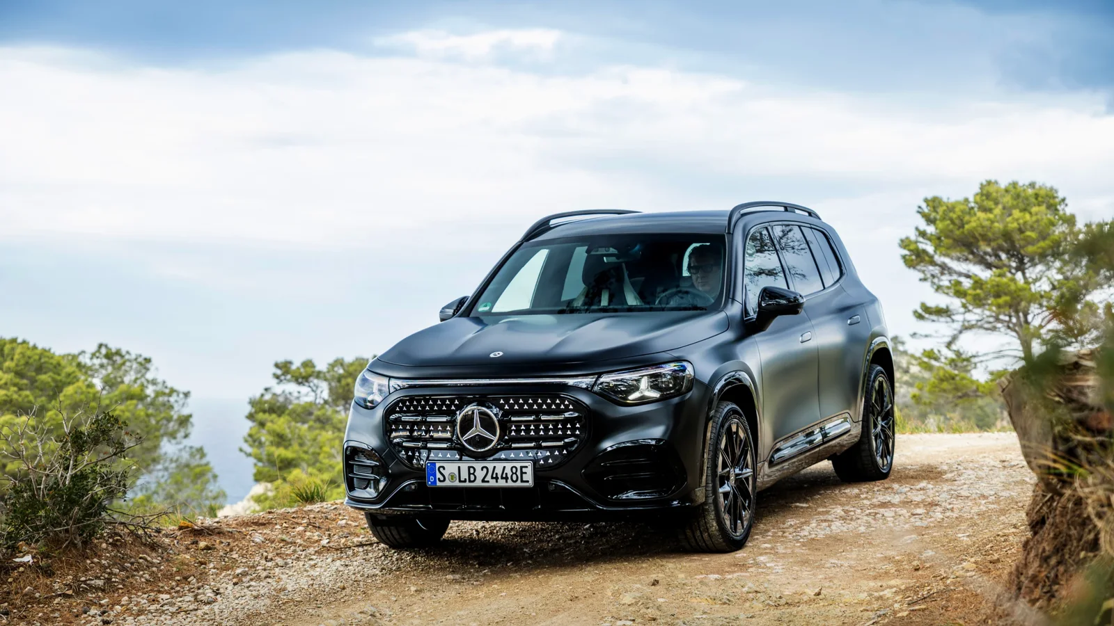 SUV Mercedes-Benz EQB negru, surprins frontal- lateral pe un drum de țară pietros, în peisaj cu cer senin și copaci verzi în fundal.
