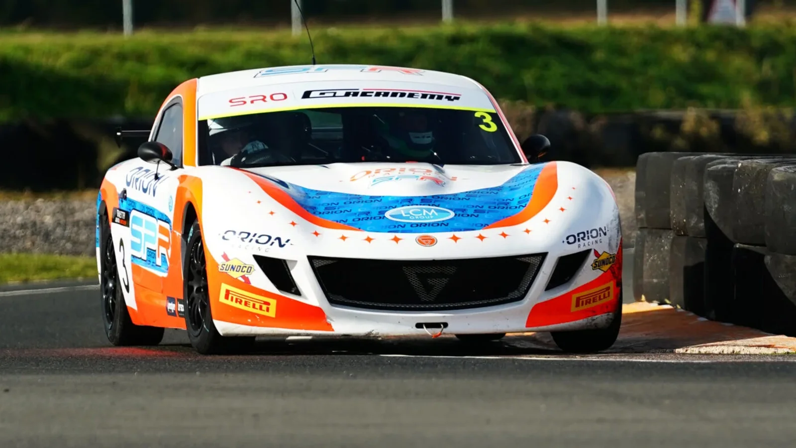Ginetta Junior albastru alb și portocaliu, fotografiat din față pe circuit, în viraj, la viteză, cu gard de protecție în dreapta.