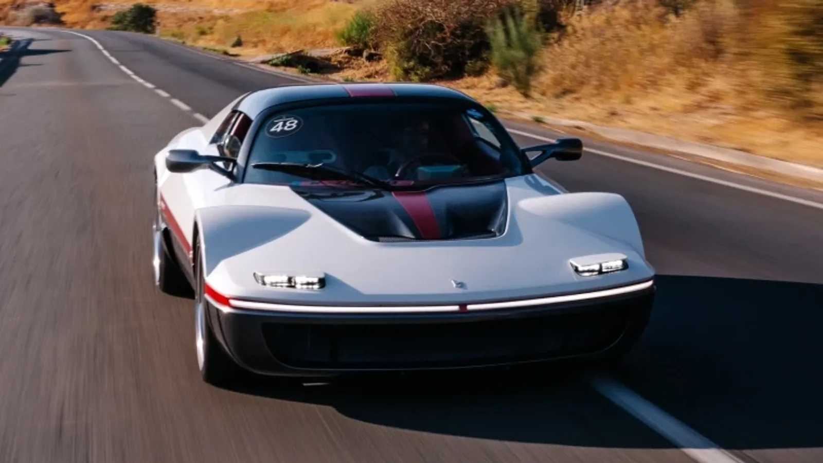 Mașina sport Bertone Runabout albă cu accente negre și roșii, văzută din față, rulând pe un drum asfaltat înconjurat de vegetație uscată și un deal stâncos pe fundal, sub cer senin, în timpul zilei.