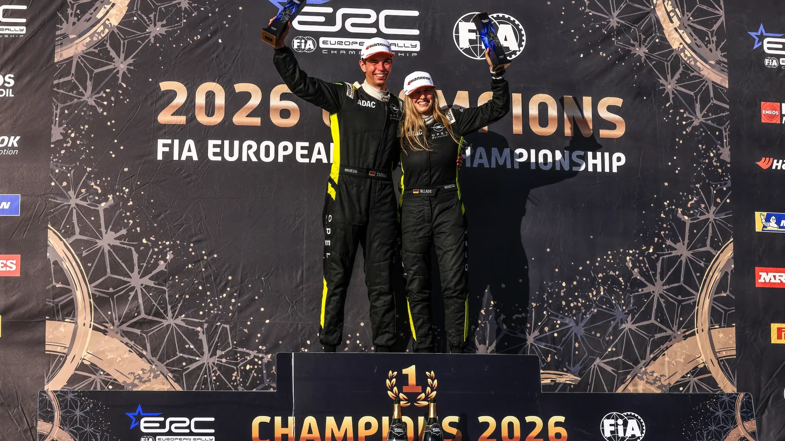 Timo Schulz și copilotul său stau pe podiumul campionilor europeni de juniori la raliuri 2026, purtând combinezoane negre. Fundalul este negru, cu text auriu „2026 FIA European Championship” și logo-uri ERC și FIA. Ambii ridică degetul arătător, celebrând victoria.
